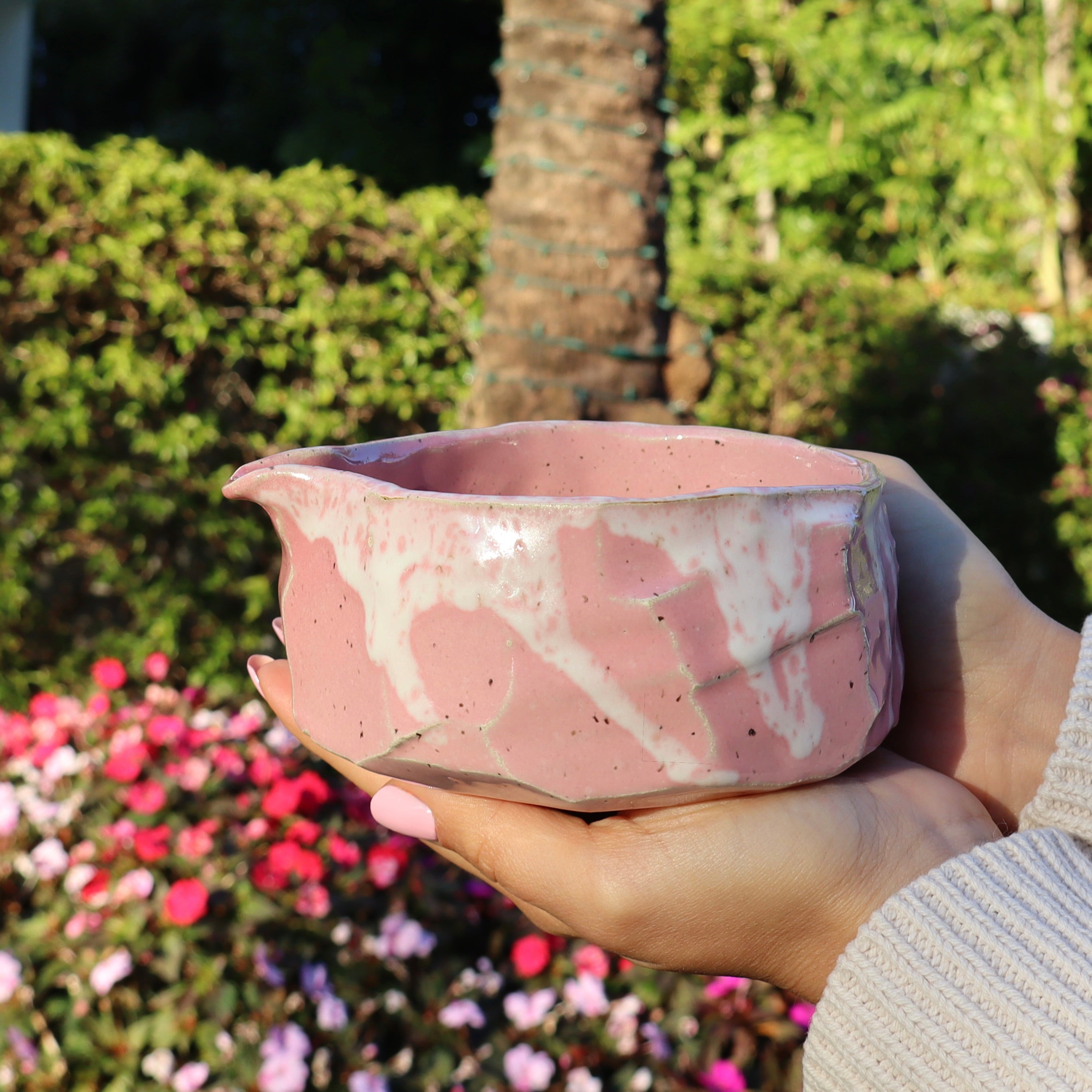 Rustic pink kurinuki chawan with pouring spout full view, held by a woman's hands in front of flowers